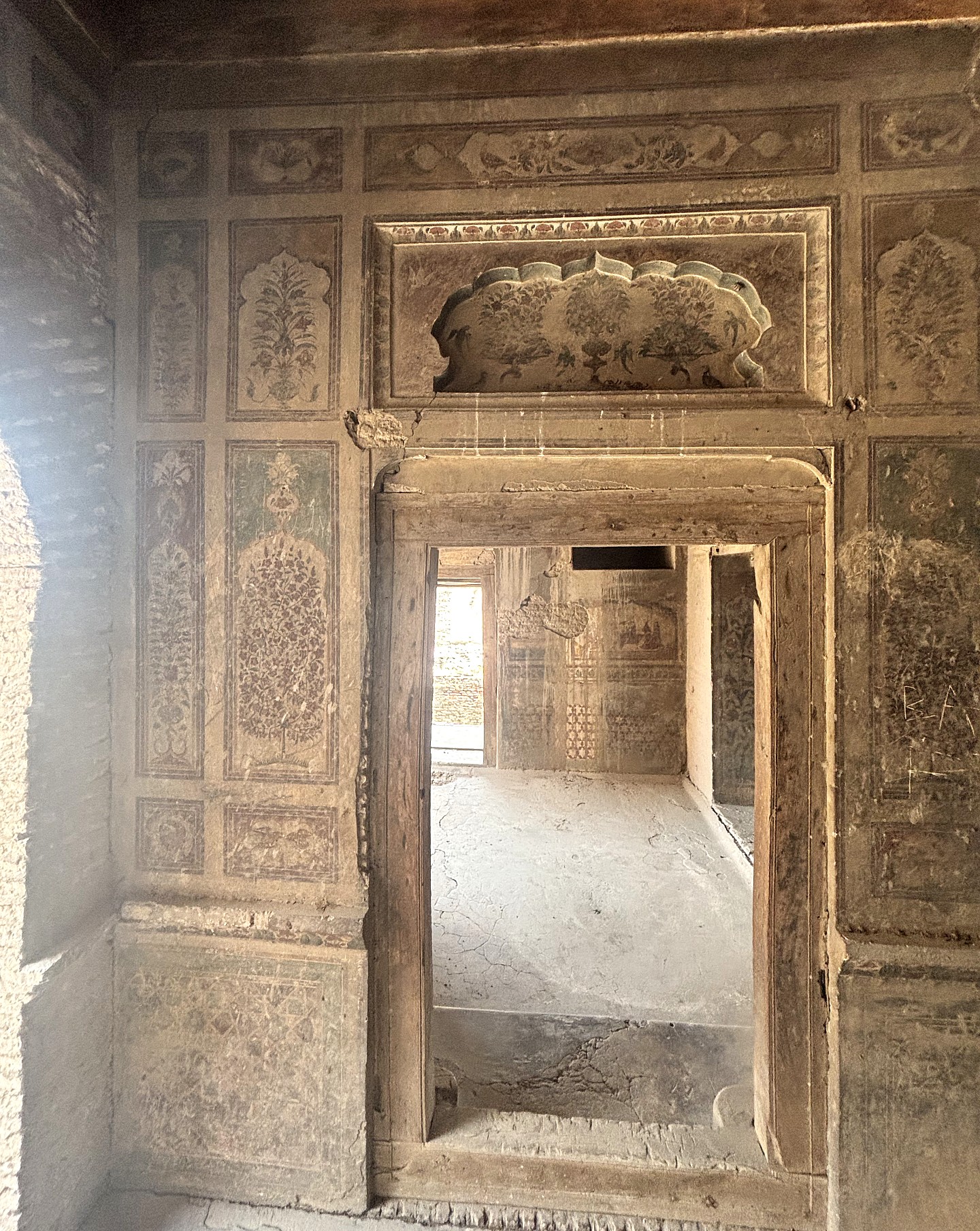 Interior of a historic building featuring faded floral wall paintings, decorative architectural details, and a wooden doorway leading into another room.