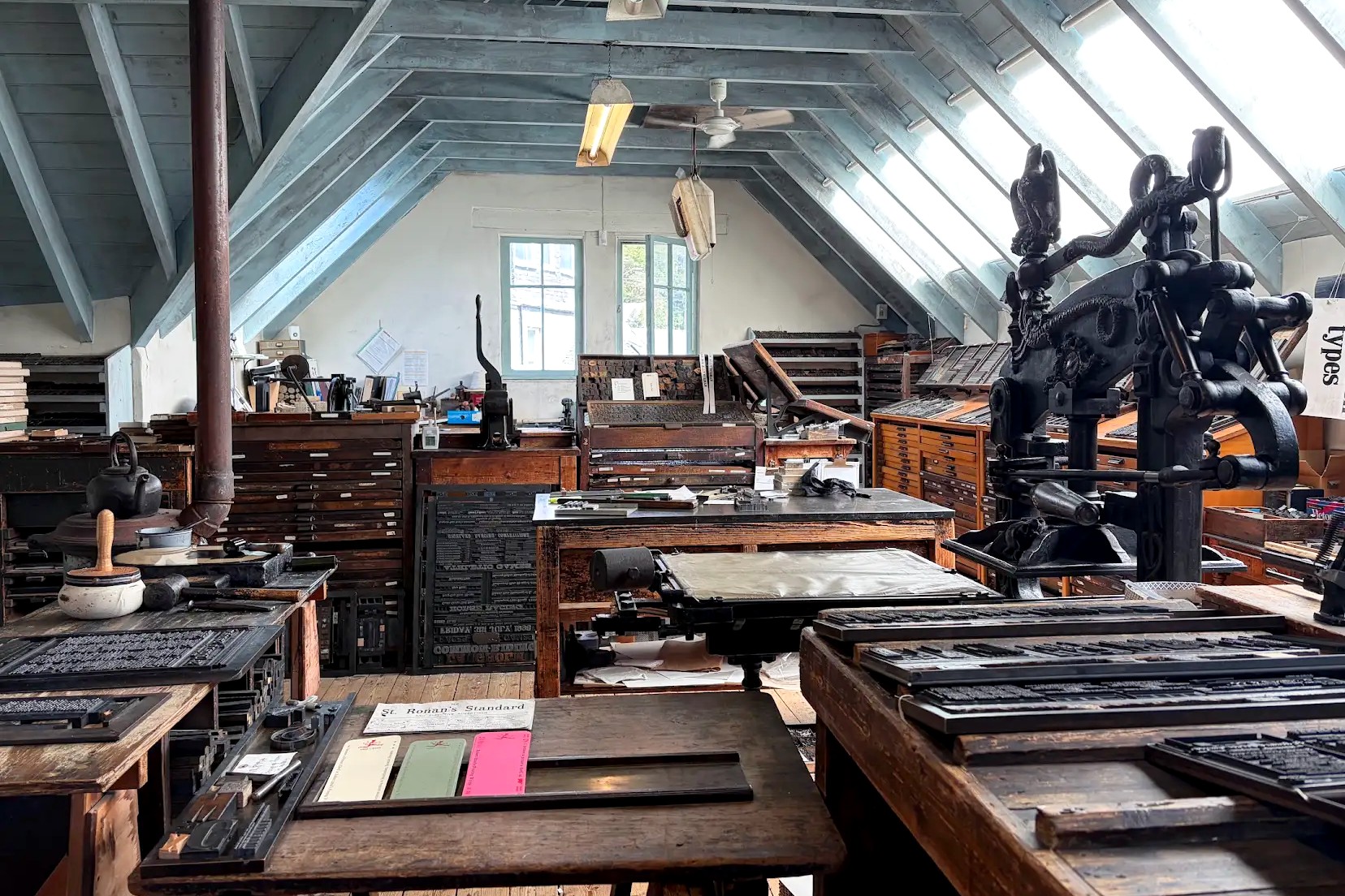 An historic letterpress printing workshop filled with antique printing presses, wooden type cabinets, trays of metal type, and worktables. Natural light streams through large slanted skylights, illuminating the traditional printmaking equipment and tools.