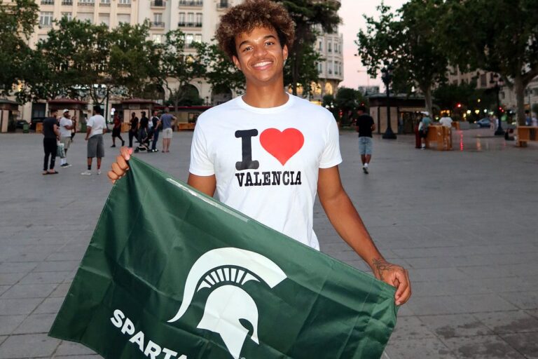 Michigan State University student Anthony Wilson smiles while holding a green MSU Spartans flag in a plaza in Valencia, Spain, wearing a white T-shirt that reads “I ❤️ Valencia,” with historic buildings in the background.