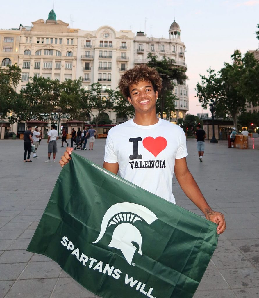 Michigan State University student Anthony Wilson smiles while holding a green MSU Spartans flag in a plaza in Valencia, Spain, wearing a white T-shirt that reads “I ❤️ Valencia,” with historic buildings in the background.