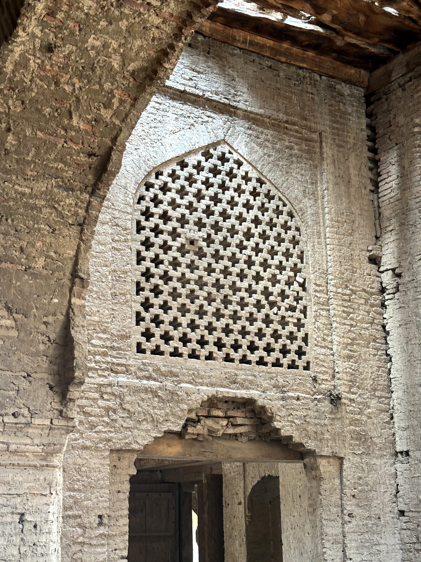 Intricate geometric brick latticework fills a pointed arch above a doorway inside a historic building, surrounded by weathered brick walls and exposed wooden ceiling beams.