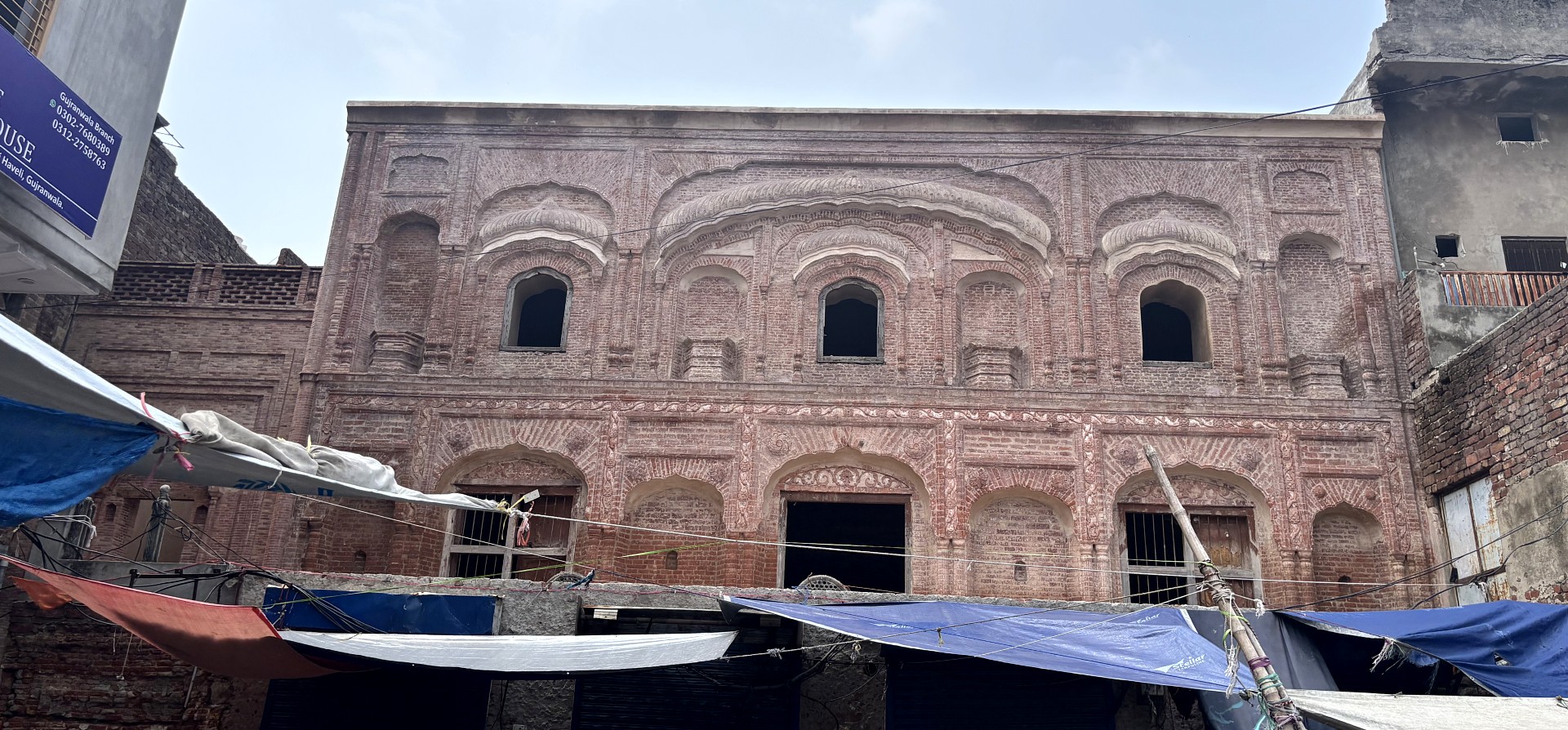 Exterior of a two-story historic brick building featuring decorative arches, recessed windows, and intricate brickwork, with market stalls and fabric awnings along the street below.