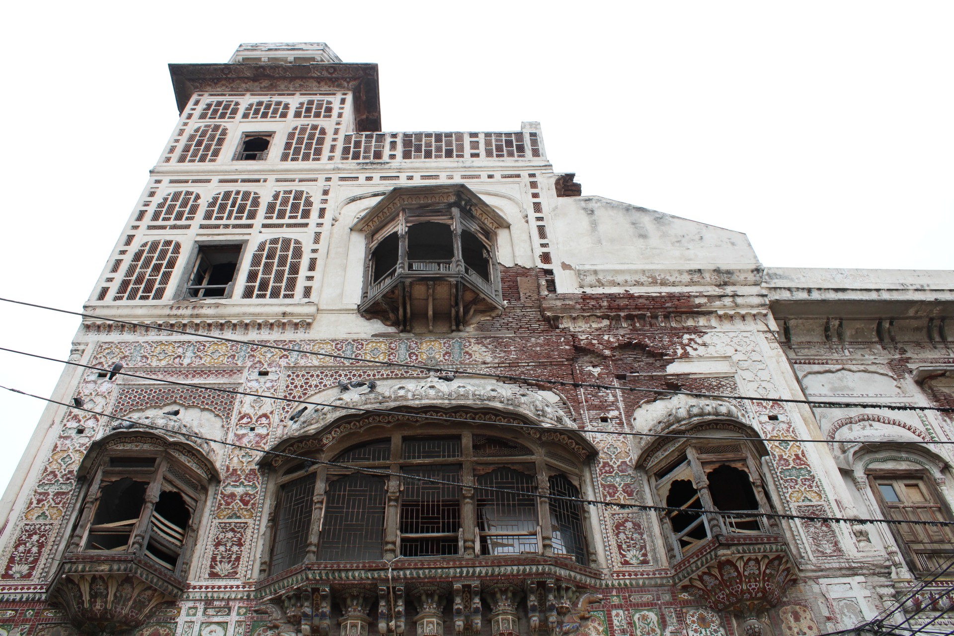 Exterior of a historic multistory building featuring elaborately decorated walls, intricate tilework, projecting wooden balconies, and lattice windows, with sections of deteriorating plaster exposing the underlying brickwork.