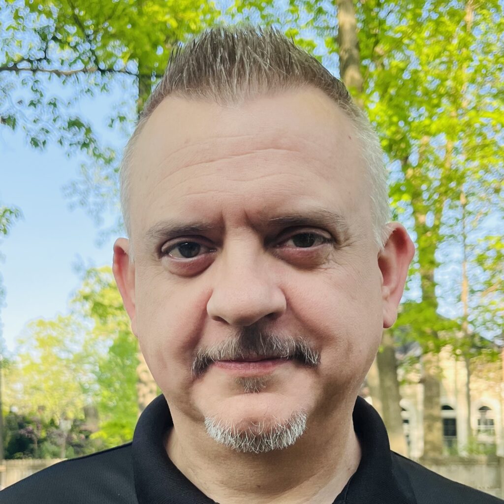 Head shot of Jesse Draper in a black polo shirt in front of some oak trees and blue sky.